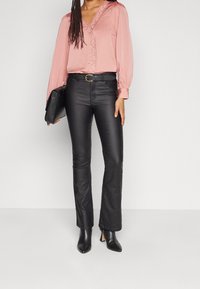 Blouse en satin rose avec bordure en dentelle, manches longues ; pantalon évasé en cuir synth étique noir. Mannequin tenant une pochette noire, portant des bottines à talons pointues.