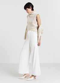 Maglione senza maniche lavorato a maglia color crema con colletto alto, abbinato a pantaloni bianchi larghi con tasche. Il modello indossa décolleté sottili di colore chiaro.