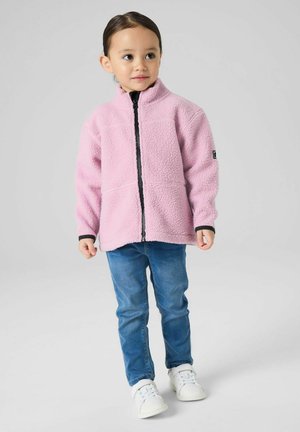Jeune enfant debout, portant une veste polaire rose, un jean bleu et des baskets blanches, regardant légèrement sur le côté.