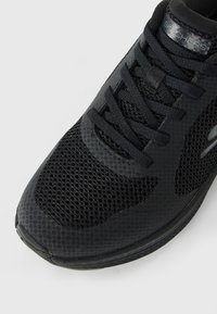 Artimas juodų Skechers sportbačių, pagamintų iš tinklelio audinio, vaizdas. Atskiros detalės – raišteliai ir logotipas ant liežuvio bei šono, priešais baltą neutralų foną.