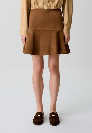 Falda acampanada de ante marrón combinada con una blusa beige de manga larga y mocasines marrón oscuro con detalles dorados sobre un fondo claro.