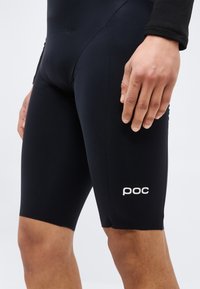 Κοντινό πλάνο ενός ατόμου που φοράει μαύρο σορτς ποδηλασίας με το λογότυπο "poc" στον μηρό, στέκεται με το ένα χέρι χαλαρό δίπλα του.