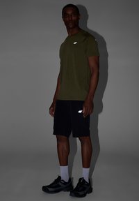 Homme debout en t-shirt vert olive et short noir avec logo blanc, portant des chaussettes blanches et des chaussures de sport noires, sur fond gris.