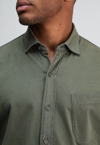 Olijfgroene overhemd met knopen, gemaakt van lichtgewicht stof, voorzien van een zak met geborduurd logo, subtiele textuur en standaard kraag.