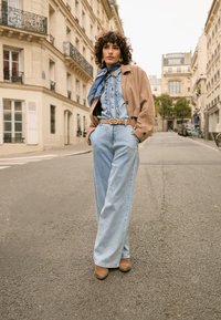 Jachetă maro din piele întoarsă peste o ținută din denim albastru deschis, cu un top cu volane și blugi cu picioare largi, accesorizată cu o eșarfă albastră și o curea maro.