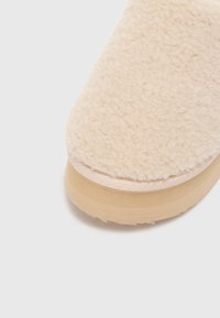 Beige, fluffy tøffel med en myk, teksturert overflate og en flat, lys brun gummisåle. Glatte kanter og en avrundet tådesign.