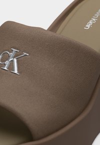 Καφέ σαγιονάρα με υφασμάτινο λουρί με υφή και ασημί λογότυπο "CK" στην κορυφή, μπεζ πέλμα και στρογγυλεμένο ανοιχτό σχέδιο στα δάχτυλα.