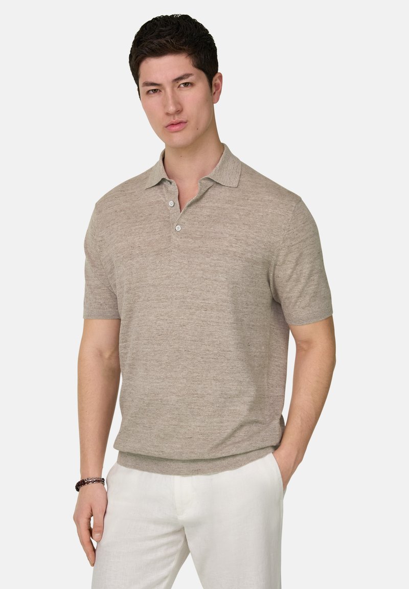 Homme portant un polo en maille beige à manches courtes et un pantalon blanc, debout avec une main dans la poche, sur un fond uni.