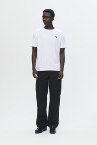 T-shirt en coton blanc avec un col rond et un petit logo noir sur la poitrine, associé à un pantalon large noir et des chaussures noires.