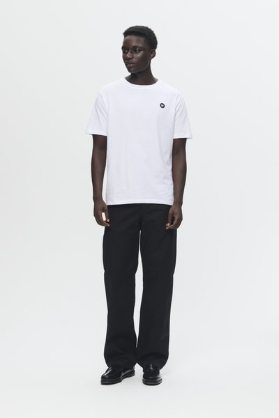 Camiseta blanca de algodón con cuello redondo y pequeño logo negro en el pecho, combinada con pantalones anchos negros y zapatos negros.
