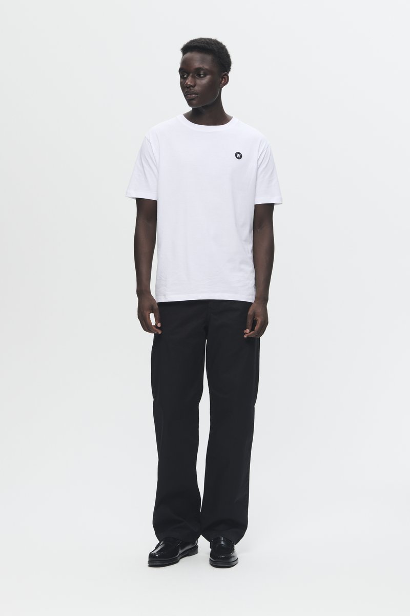 T-shirt en coton blanc avec un col rond et un petit logo noir sur la poitrine, associé à un pantalon large noir et des chaussures noires.