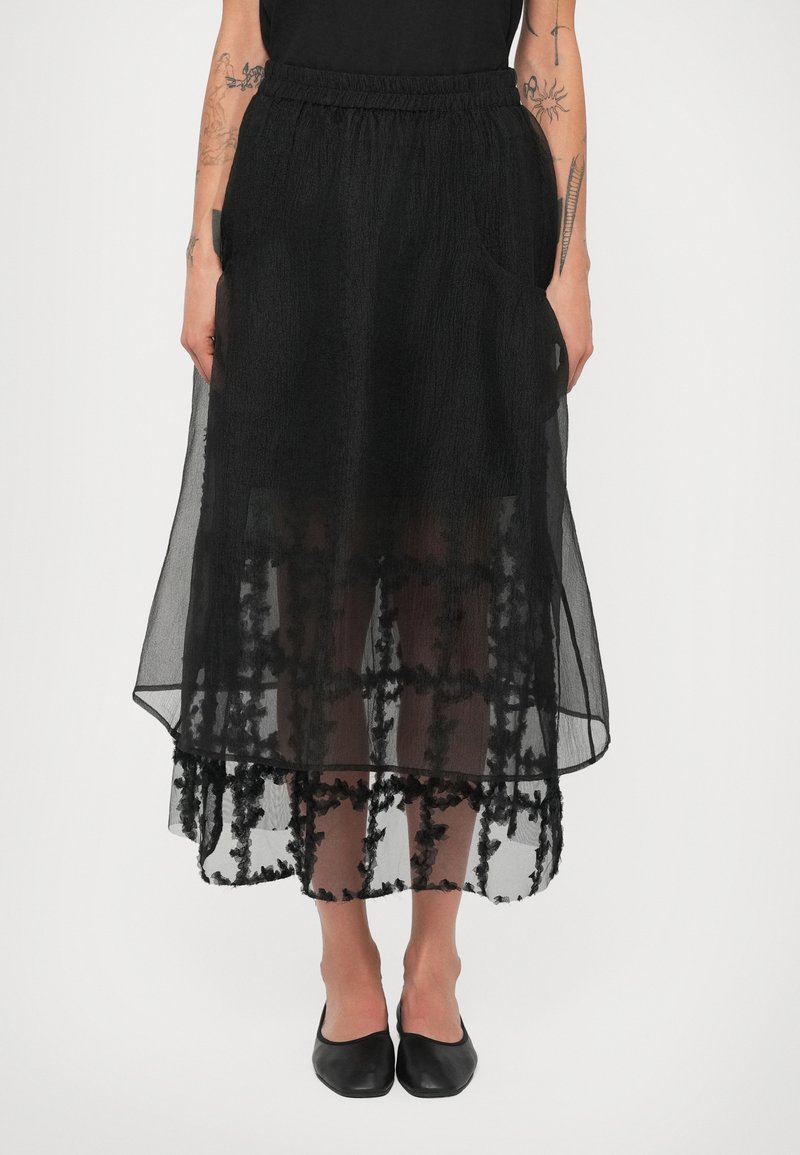 Melnā caurspīdīgā svārku ar tekstūru, kas raksturojas ar slāņainu dizainu un ziedu motīvu apakšmalā; elastīga josta un sānu kabatas.