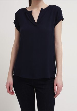 Vrouw draagt een marineblauwe korte mouwen blouse met een V-hals en zwarte broek, staand tegen een effen lichte achtergrond.