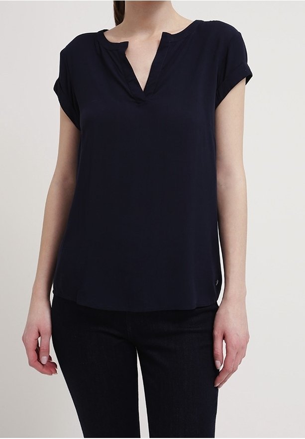Femme portant un chemisier bleu marine à manches courtes avec un col en V entaillé et un pantalon noir, debout devant un fond clair uni.