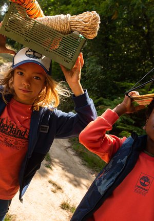 Deux enfants transportent une caisse verte à l'aide d'une corde. Ils portent des vêtements décontractés : un t-shirt rouge Timberland, des vestes bleue et rouge, et une casquette sombre.