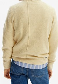 Pull en polaire de couleur crème avec une surface texturée, doté d'une couture verticale et d'une surpiqûre décorative près de l'ourlet, superposé à une chemise à rayures bleues.