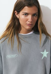 Sweat à capuche gris avec un col rond, présenté avec une étoile verte menthe et un texte indiquant "SHINE BRIGHT Browinie Original" dans un label arrondi.