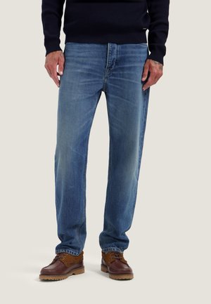 Homme portant un jean bleu droit, des bottes en cuir marron et un haut sombre à manches longues, debout avec les mains détendues le long du corps.