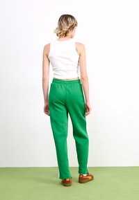 Grønne sweatpants med hvide sidestropper, elastisk talje og løs pasform, parret med en tætsiddende hvid tanktop og brune sneakers.