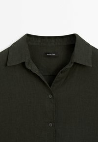 Chemise boutonnée en lin vert foncé avec col camp, présentant une texture douce et de subtils détails de couture. L'étiquette indique "Massimo Dutti."