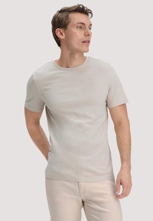 Young man in light beige t-shirt and pants stands with one hand behind back, looking to the side against plain background.