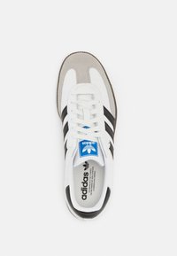 Zapatillas Adidas con un upper de cuero blanco, acentos de gamuza gris, tres rayas negras y una etiqueta de logo azul en la lengüeta.