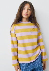 Jeune fille avec de longs cheveux bruns portant un pull rayé jaune et lavande et un jean bleu avec des motifs circulaires, se tenant devant un mur blanc.