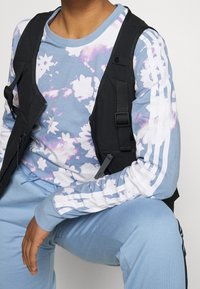 Black zip-up vest over a light blue long-sleeve shirt with purple floral patterns. Light blue sweatpants with ribbed texture and side stripes.