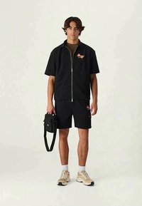 Svart kortärmad zip-tröja med logotyp, matchande shorts, beige sneakers och en svart axelremsväska. Slät textur, minimalistisk design.