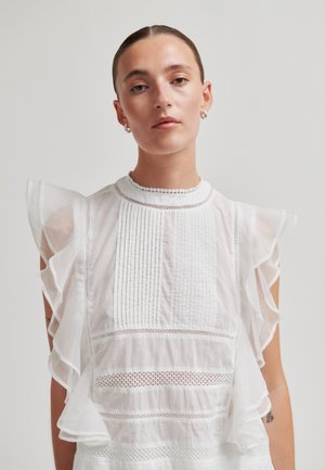 Kvinne med bakoverglattet hår som har på seg en hvit, gjennomsiktig bluse med volangerm og blonde detaljer på en ensfarget bakgrunn.