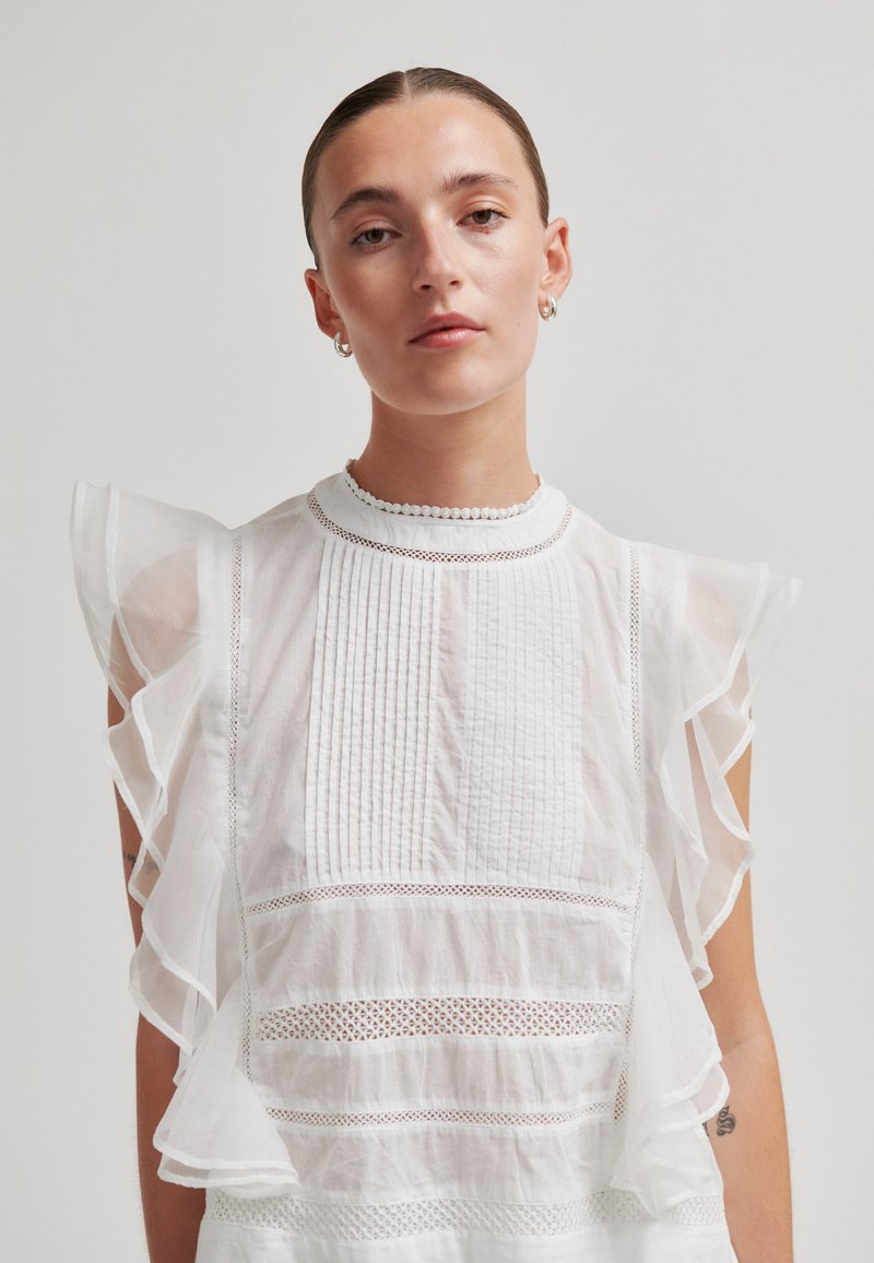 Femme aux cheveux plaqués en arrière portant un chemisier blanc transparent avec manches à volants et détails en dentelle sur un fond uni.
