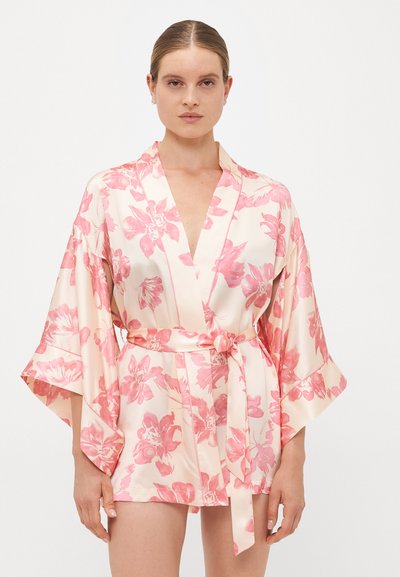Femme portant un peignoir court en soie crème avec des motifs floraux roses, des manches larges et une ceinture nouée à la taille, se tenant devant un fond uni.