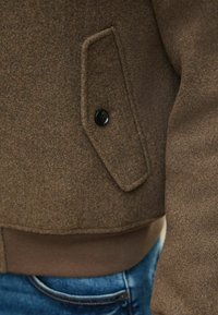 Jachetă din lână maro cu tiv ribbed maro, buzunar lateral cu închidere buton și țesătură texturată. Blugi albaștri vizibili în partea de jos.