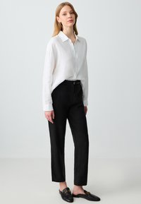 Weißes Leinenhemd mit Knopfleiste, schwarze hochtaillierte Hose und schwarze Slip-On-Schuhe mit goldenen Akzenten.