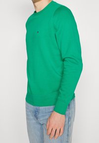 Felpa verde realizzata in tessuto testurizzato con colletto rotondo e maniche lunghe, dotata di un piccolo logo sul petto, abbinata a jeans in denim chiaro.