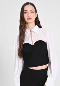Top corto nero con scollatura a cuore, abbinato a una blusa bianca trasparente a maniche lunghe con apertura a chiave e bottone argentato.