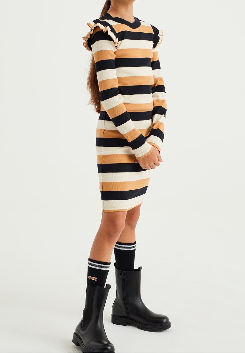 Enfant portant une robe à rayures beige, noire et blanche avec des épaules à volants, des bottes noires et des chaussettes noires montantes à rayures blanches.