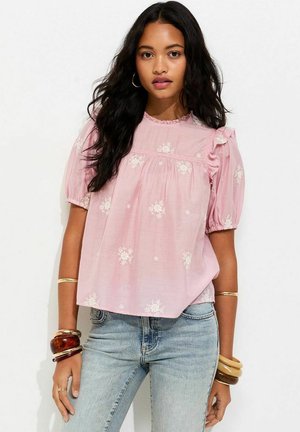 Młoda kobieta o długich ciemnych włosach nosi różową kwiecistą bluzkę z bufkami i jasnoniebieskie dżinsy, udekorowana bransoletkami i kolczykami kołami.