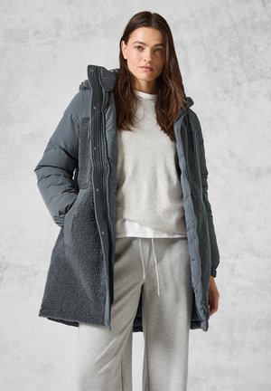 Manteau d'hiver long et gris foncé avec un extérieur texturé et pelucheux. Équipé d'une capuche, d'une fermeture éclair et de poches latérales ; porté avec un pull gris clair et un pantalon.