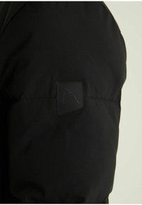 Schwarze gepolsterte Jacke aus strukturiertem Stoff, mit einem quadratischen Logo-Patch am Ärmel und einer glatten Oberfläche.