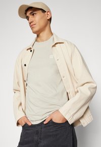 bežová džínová bunda s čiernymi gombíkmi, svetlošedé tričko s malým logom a tmavosivé džínsy, doplnené zodpovedajúcou bežovou šiltovkou.