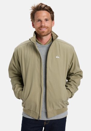 Hombre con cabello y barba castaño claro, vistiendo una chaqueta beige con cremallera sobre un suéter gris, con las manos en los bolsillos de la chaqueta, sonriendo levemente.