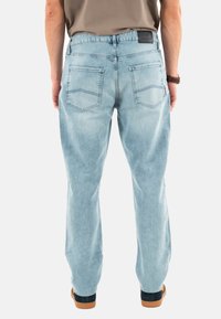 Pantalones de mezclilla azul claro con un corte relajado, bolsillos traseros visibles y detalles cosidos. Llevados con zapatos marrones y una camisa beige.