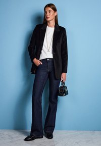 Blazer noir à finition lisse, associé à un haut blanc et un jean en denim foncé évasé. Les accessoires comprennent un petit sac à main noir et un collier en or.