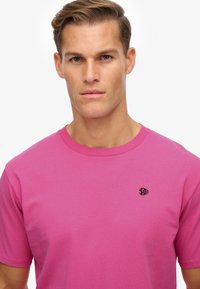 Camiseta de algodón rosa con cuello redondo, mangas cortas y un pequeño logo negro en el lado izquierdo del pecho. Color sólido, textura suave.