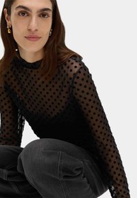 Svart nättröja med långa ärmar och prickigt mönster, med hög halsringning och svart underlayer. Matcher med mörkgrå denimbyxor.