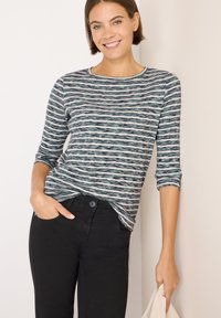 Maglietta a maniche lunghe a righe nei toni del verde, nero e bianco; tessuto morbido; scollo rotondo; indossata con pantaloni neri; vestibilità casual.