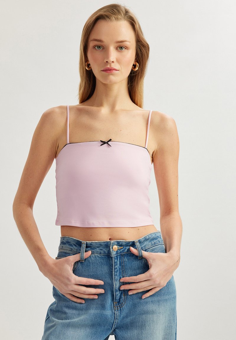 Lyserød kort topp med tynde stropper, der har en sort kant og en lille sort sløjfe i midten. Bæres med højtaljede denim jeans.