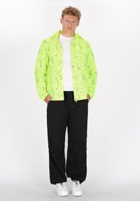 Neon gul jakke med abstrakt sort mønster, frontlynlås og hætte, parret med sorte bukser og hvide sneakers. Klassisk pasform.