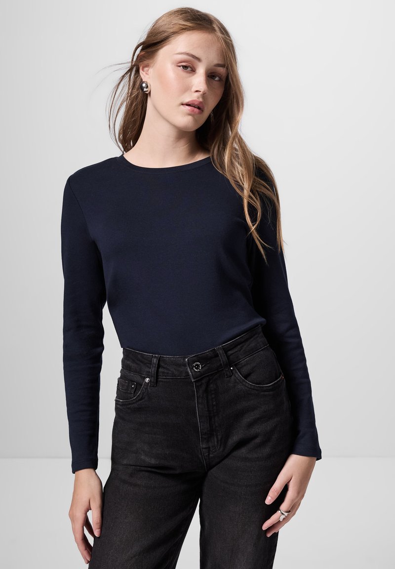 Marineblauw langemouwen top gecombineerd met hoge zwarte jeans. De top heeft een ronde halslijn en de jeans hebben een knoopsluiting en voorzakken.